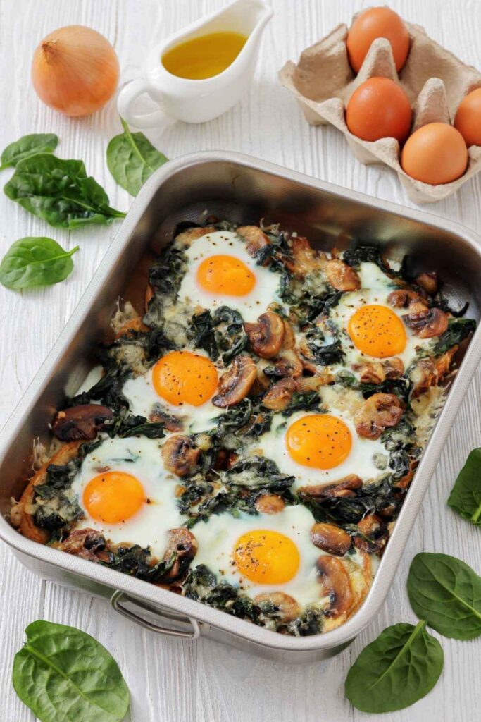A baking dish with cooked eggs, mushrooms, and spinach sits on a white table surrounded by fresh spinach leaves, eggs in a carton, an onion, and a small pitcher of oil.
