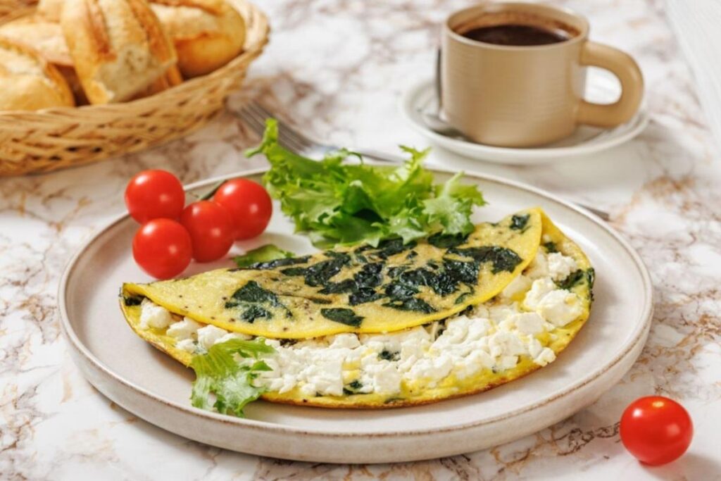 A plate with a spinach and cheese omelette, cherry tomatoes, and lettuce, alongside a cup of coffee and a basket of bread on a marble table.