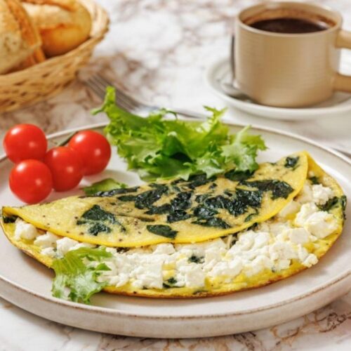 A plate with a spinach and cheese omelette, cherry tomatoes, and lettuce, alongside a cup of coffee and a basket of bread on a marble table.