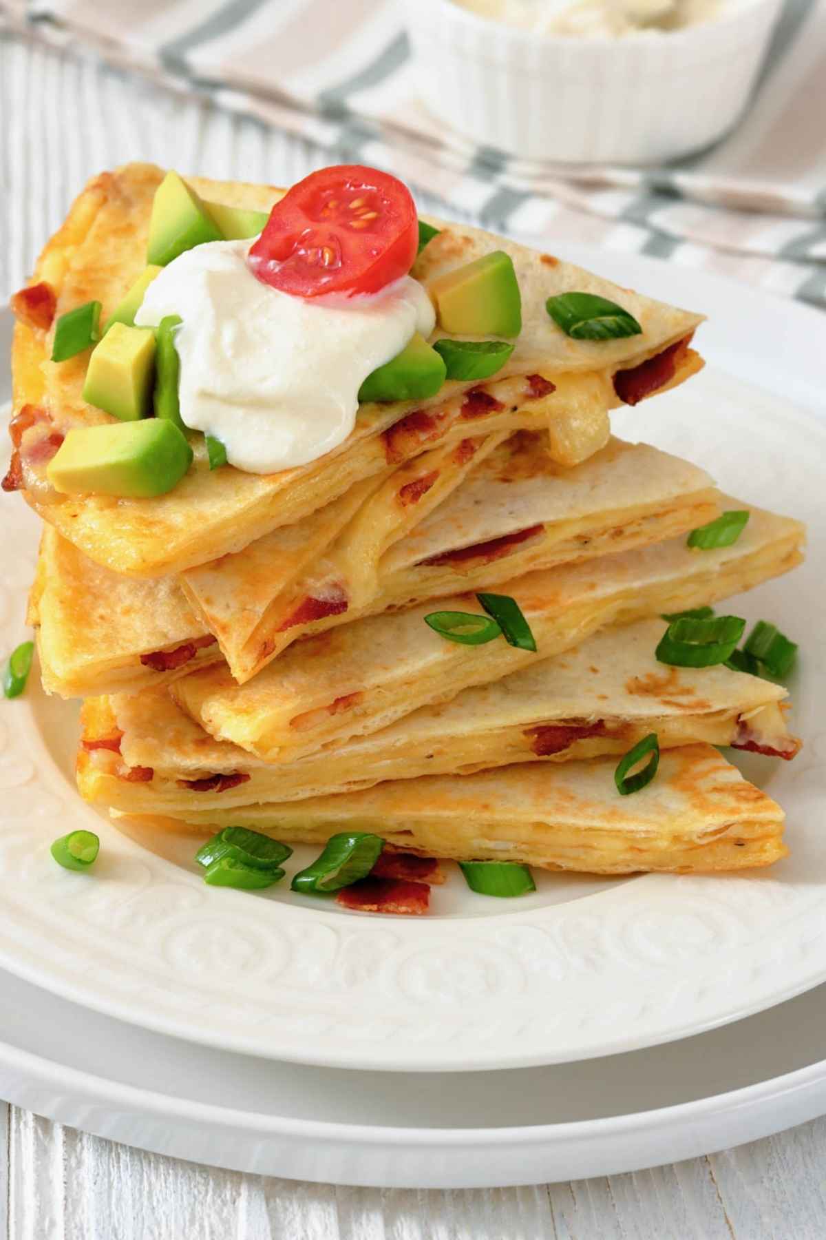 A stack of quesadilla wedges topped with sour cream, diced avocado, green onions, and a cherry tomato slice on a white plate.