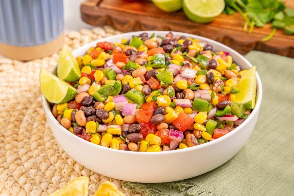 A white bowl filled with a colorful mix of beans, corn, diced tomatoes, onions, and peppers, garnished with lime wedges.
