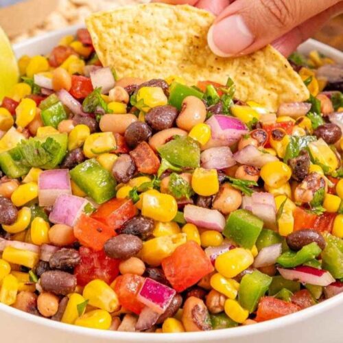 A bowl of Cowboy Caviar garnished with lime wedges. A hand is dipping a tortilla chip into the salad.