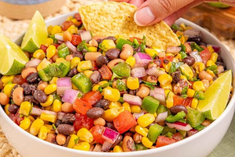 A bowl of Cowboy Caviar garnished with lime wedges. A hand is dipping a tortilla chip into the salad.