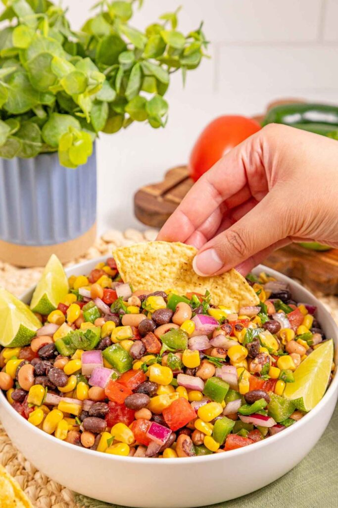 A hand dips a tortilla chip into a bowl of colorful bean and corn salad garnished with lime wedges, with a plant and vegetables in the background.