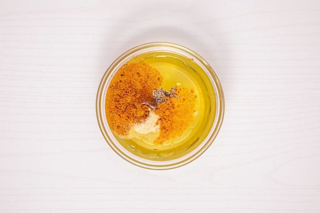 A glass bowl containing oil, ground spices, and seasoning powders sits on a light-colored wooden surface.