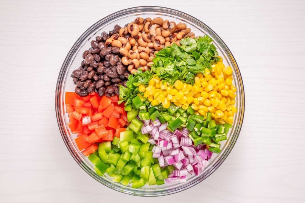 A glass bowl containing black beans, black-eyed peas, chopped cilantro, corn, red bell pepper, green bell pepper, red onion, and jalape&ntilde;o, arranged in sections on a white surface.