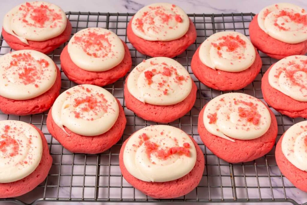 A cooling rack with rows of pink cookies topped with white frosting and pink sprinkles, placed on a marble surface.