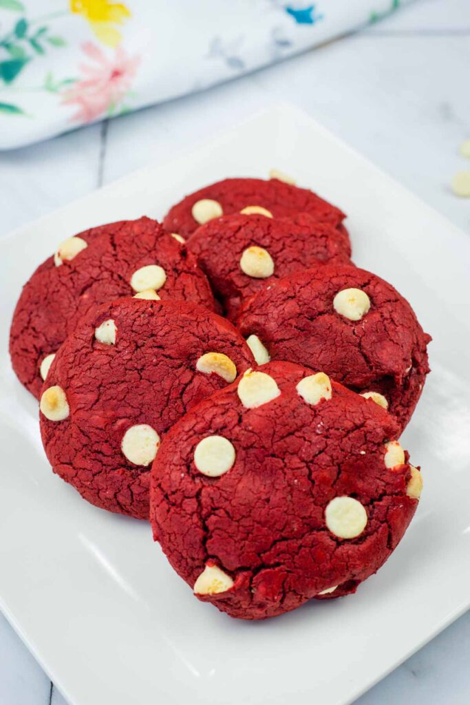 Six red velvet cookies with white chocolate chips arranged on a white square plate.