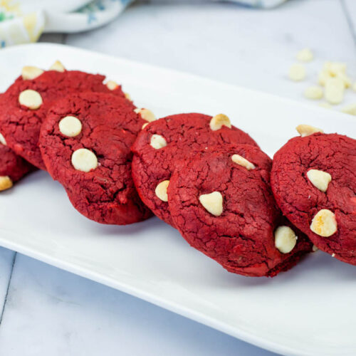 Five red cookies with white chocolate chips arranged in a row on a rectangular white plate, with a marble countertop background.