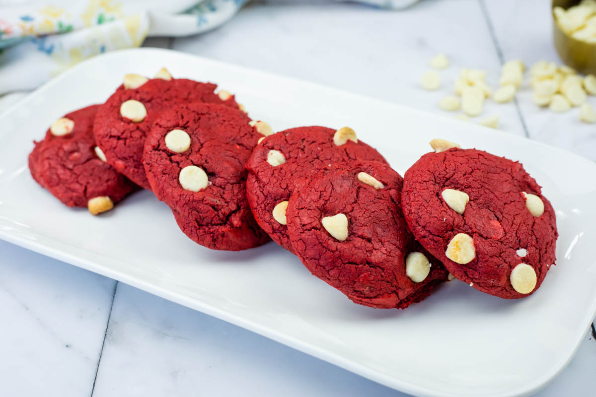 Five red cookies with white chocolate chips arranged in a row on a rectangular white plate, with a marble countertop background.