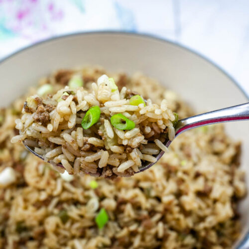 A close-up of a spoon holding dirty fried rice mixed with ground meat and sliced green onions above a pot of the same dish.