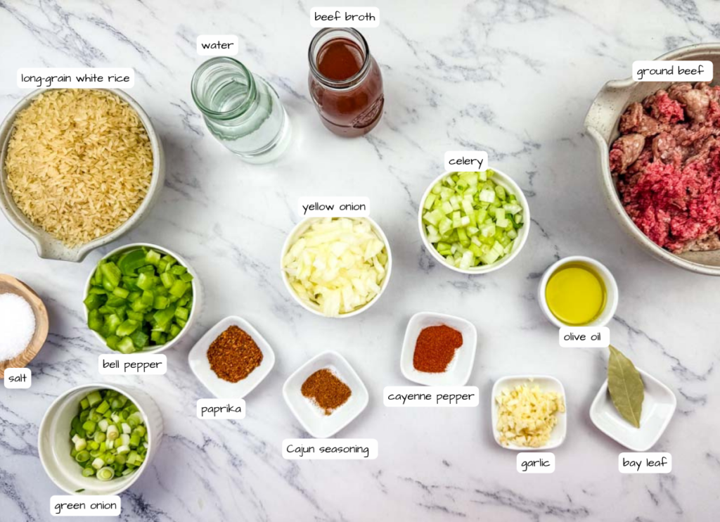 Ingredients for a beef and rice dish, including ground beef, long-grain white rice, chopped vegetables, spices, olive oil, beef broth, and water, arranged on a marble countertop.