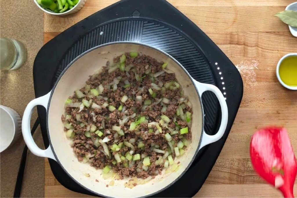 A white pot on a stovetop contains ground meat, chopped onions, and green bell peppers being cooked. Various ingredients are arranged on the wooden counter nearby.