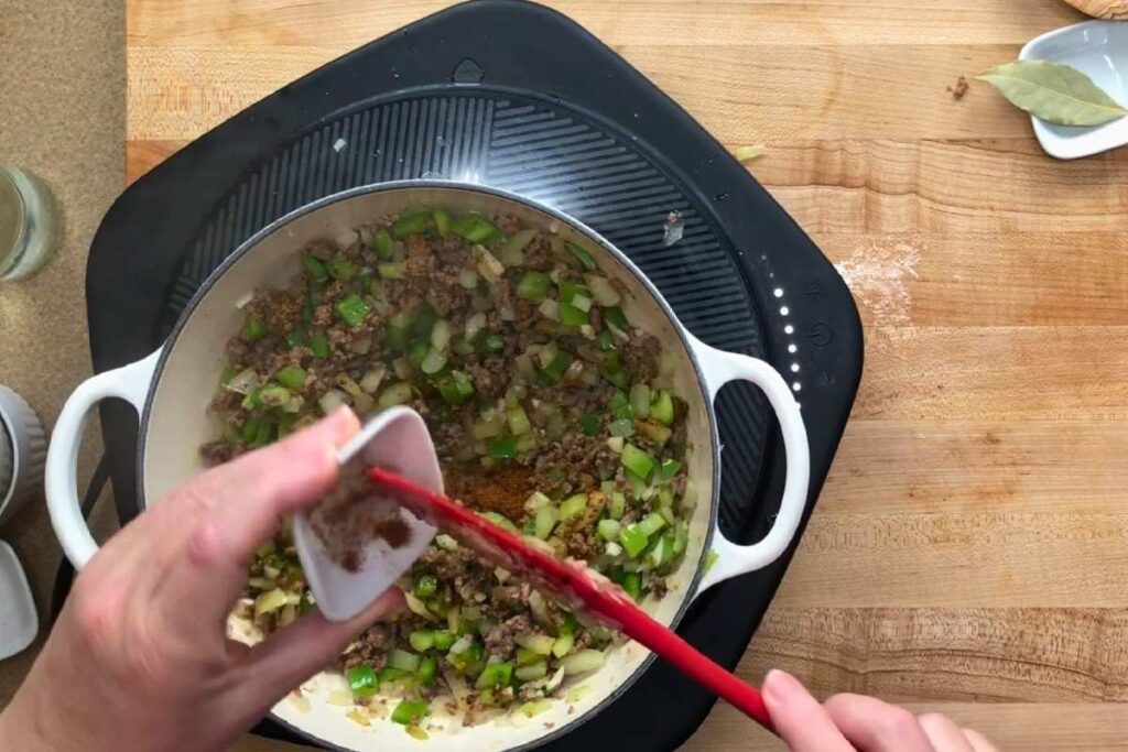 Person adds spices to a pot of ground meat, chopped onions, and green bell peppers on a stovetop. A bay leaf and utensils are on the wooden countertop nearby.