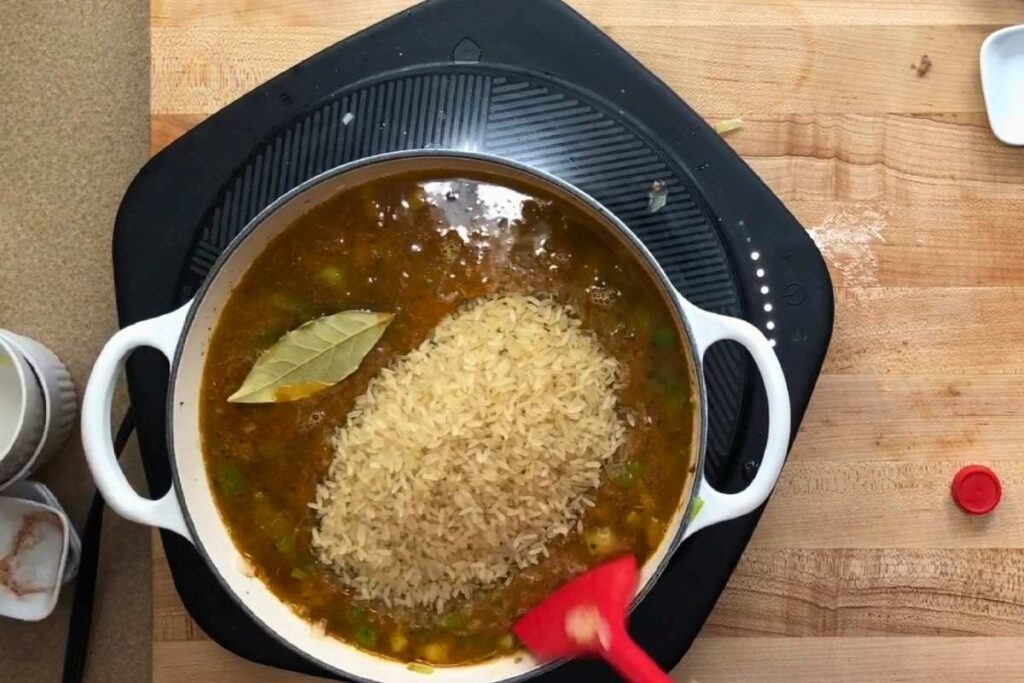 A pot with uncooked rice, bay leaf, and broth sits on an induction cooktop; a red spatula is stirring the mixture on a wooden counter.