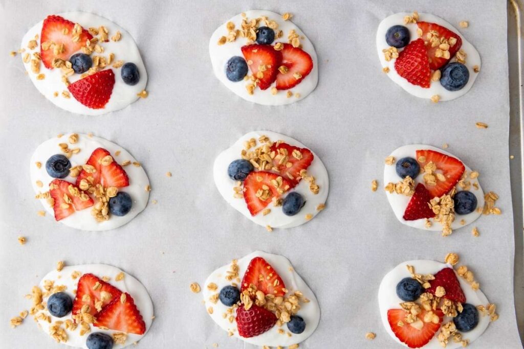 Nine oval-shaped yogurt bites topped with sliced strawberries, blueberries, and granola are arranged on a baking sheet lined with parchment paper.