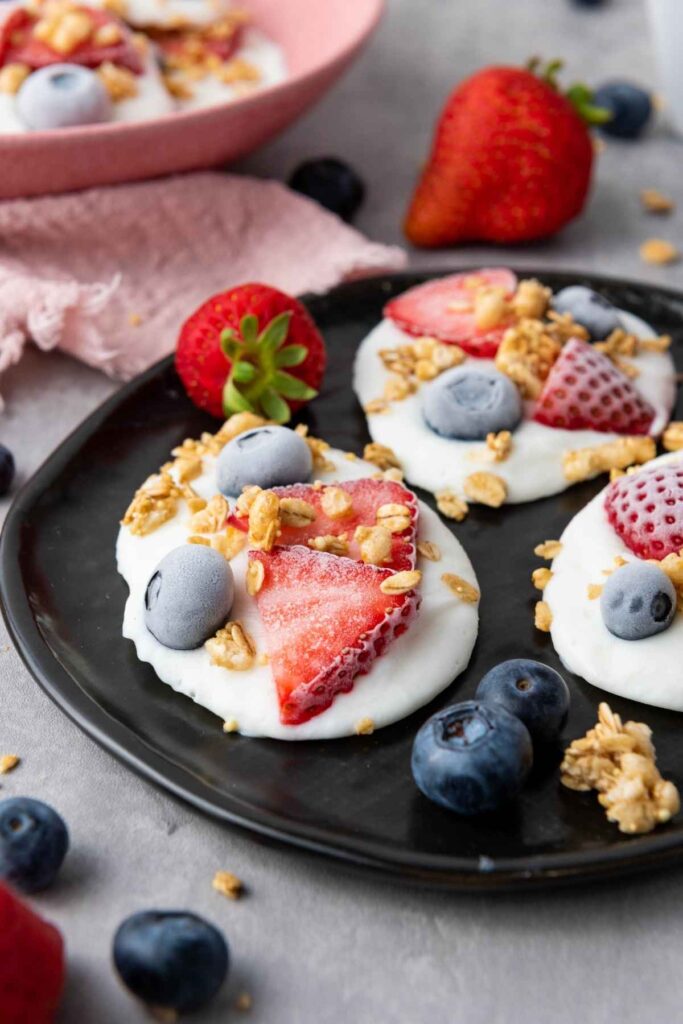 Three yogurt clusters topped with strawberries, blueberries, and granola are arranged on a black plate, with extra fruit scattered around on the table.