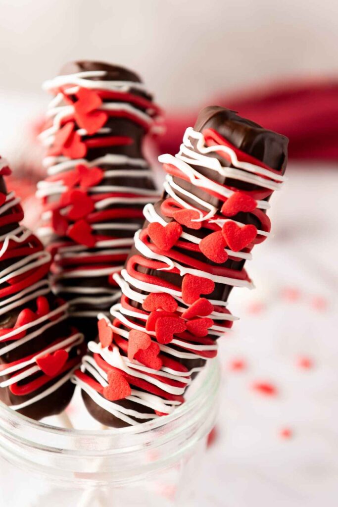 chocolate-covered marshmallow pop decorated with white icing and red heart-shaped sprinkles, displayed upright in a glass jar.