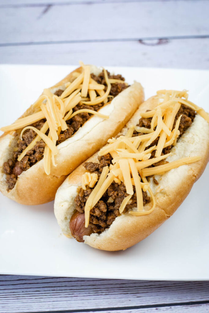 Two hot dogs on buns topped with ground meat and shredded cheddar cheese, served on a white plate.