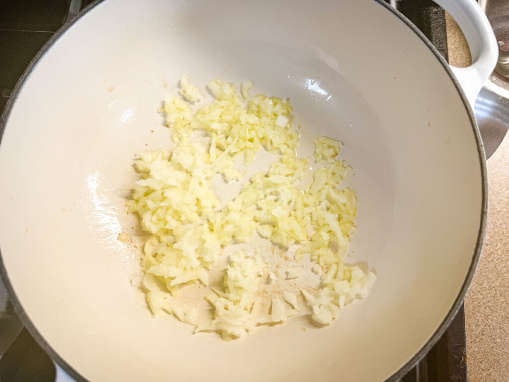 Chopped onions are being saut&eacute;ed in a white pot on a stovetop.