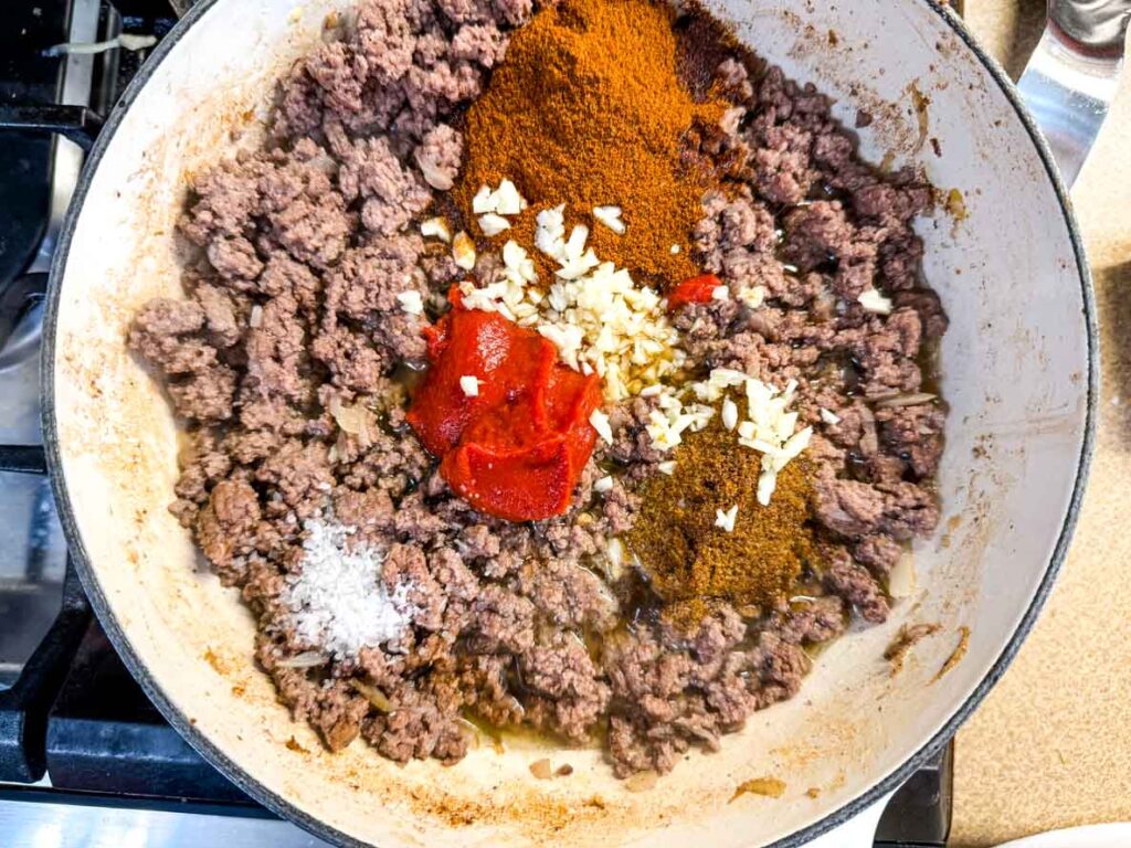 Ground beef cooking in a pan with chopped garlic, tomato paste, chili powder, and salt added on top, ready to be mixed.