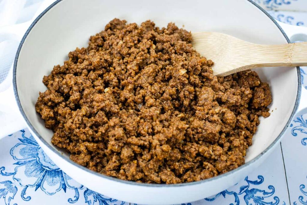 Cooked ground beef with taco seasoning in a white skillet, stirred with a wooden spatula, on a blue-patterned surface.