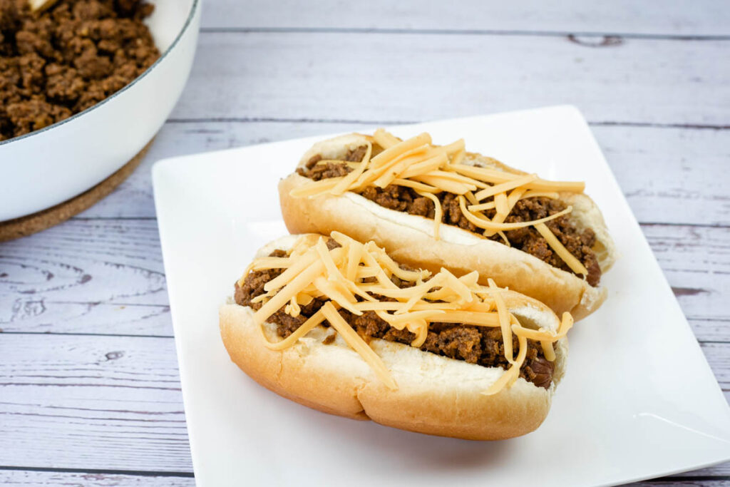 Two hot dog buns filled with ground beef and topped with shredded cheese are placed on a white square plate, with more ground beef in a dish in the background.