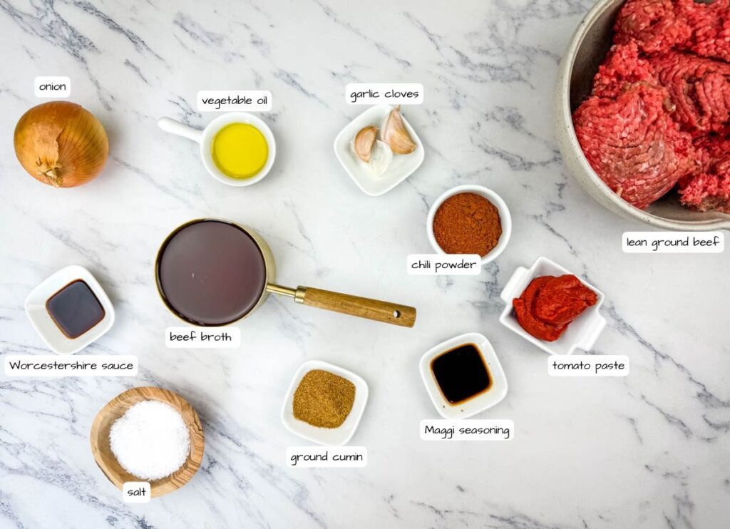 Ingredients for chili displayed on a marble surface, including ground beef, onion, garlic, vegetable oil, chili powder, tomato paste, beef broth, cumin, Maggi seasoning, Worcestershire sauce, and salt.