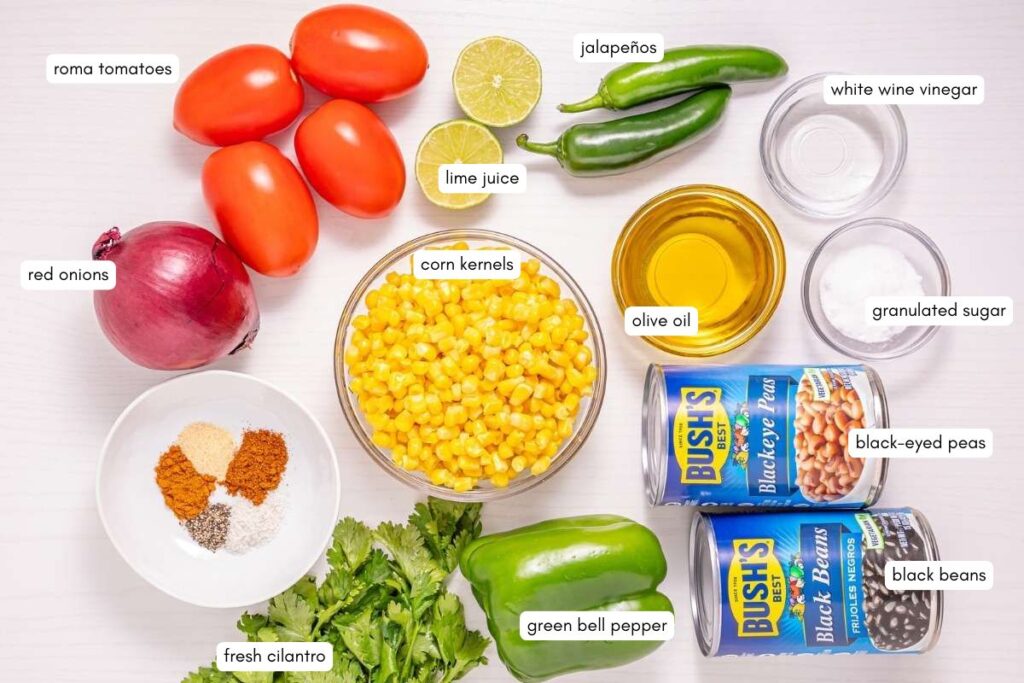 A variety of labeled ingredients for a recipe, including roma tomatoes, red onion, corn, jalape&ntilde;os, lime, olive oil, spices, fresh cilantro, green bell pepper, black-eyed peas, and black beans.