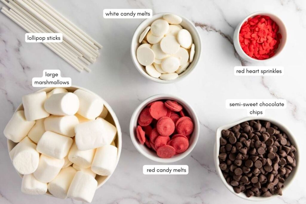 Lollipop sticks, large marshmallows, white and red candy melts, semi-sweet chocolate chips, and red heart sprinkles arranged in bowls on a marble surface.