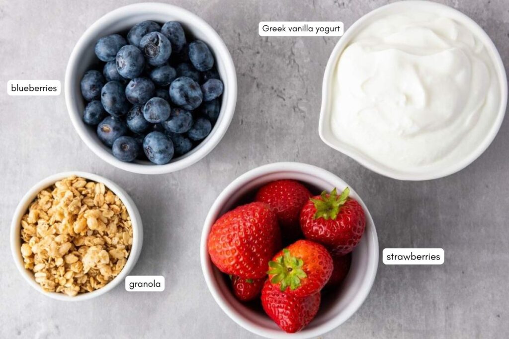 Four bowls on a gray surface containing blueberries, Greek vanilla yogurt, granola, and strawberries, each labeled with text.