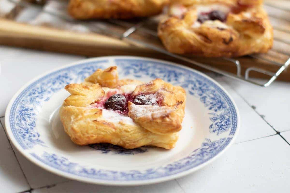 A cherry Danish pastry with powdered sugar sits on a white plate with a blue floral pattern; more pastries are on a cooling rack in the background.