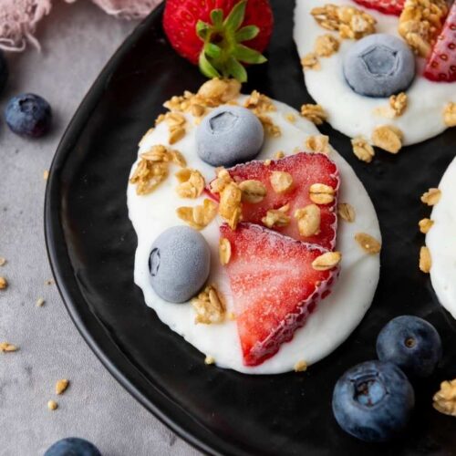 Three Frozen Vanilla Yogurt Bites topped with sliced strawberries, blueberries, and granola are arranged on a black plate, with extra berries and granola scattered around on the gray surface.