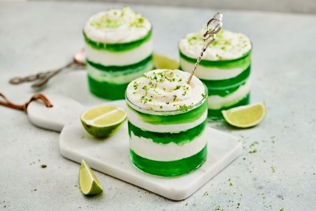 Three glasses with layered green jelly and white cream are topped with grated lime zest, placed on a white board with lime wedges beside them.