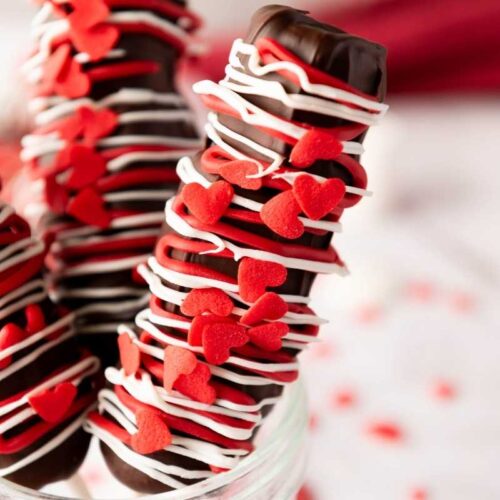 Three Valentine&rsquo;s Day Marshmallow Pops decorated with white icing and red heart-shaped sprinkles stand upright in a glass jar.