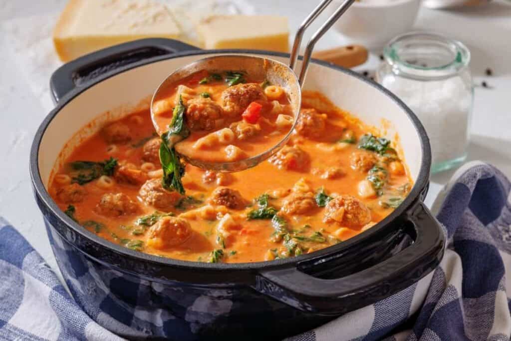 A ladle lifts meatball soup with spinach, pasta, and tomato broth from a black pot on a kitchen counter.
