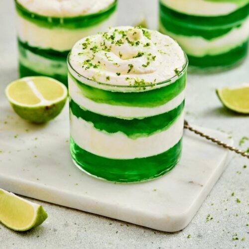 A glass dessert of St. Patrick’s Day Green Jello Parfaits, sits on a marble board with lime wedges nearby.