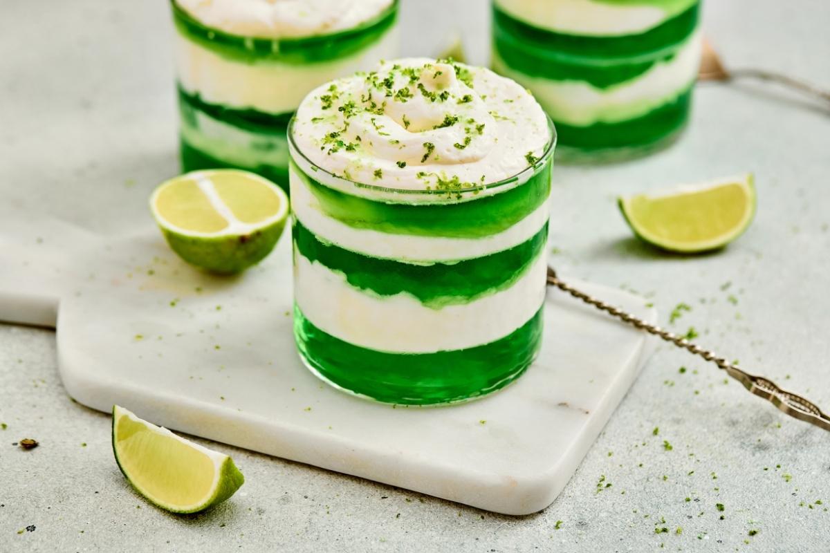 A glass dessert of St. Patrick&rsquo;s Day Green Jello Parfaits, sits on a marble board with lime wedges nearby.