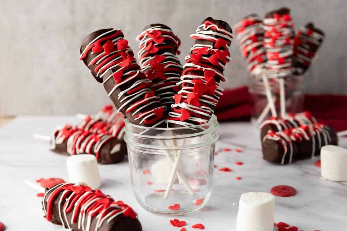 Valentine&rsquo;s Day Marshmallow Pops on sticks, decorated with red heart sprinkles and white drizzle, displayed in glass jars with loose marshmallows and sprinkles around.