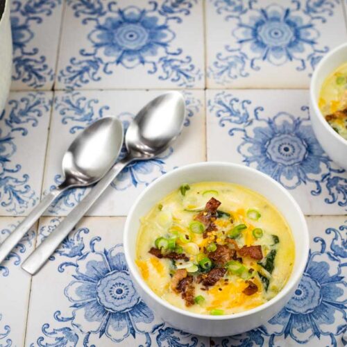 Two bowls of soup garnished with bacon and green onions sit on a decorative blue and white tiled surface next to two silver spoons and a white pot.