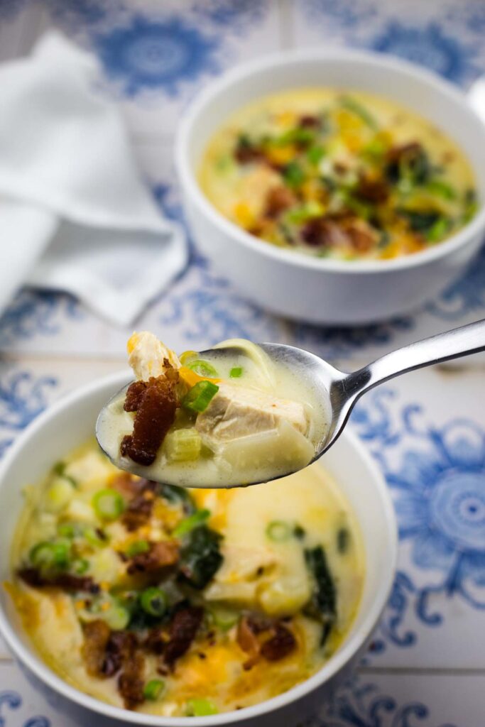 A close-up of a spoonful of creamy soup with chicken, bacon, and green onions held above a bowl, with another bowl of soup in the background.