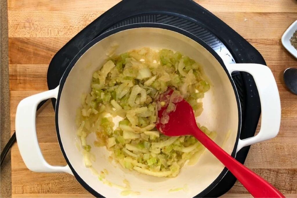 A red spatula stirs chopped onions and celery cooking in a white pot on a wooden countertop.