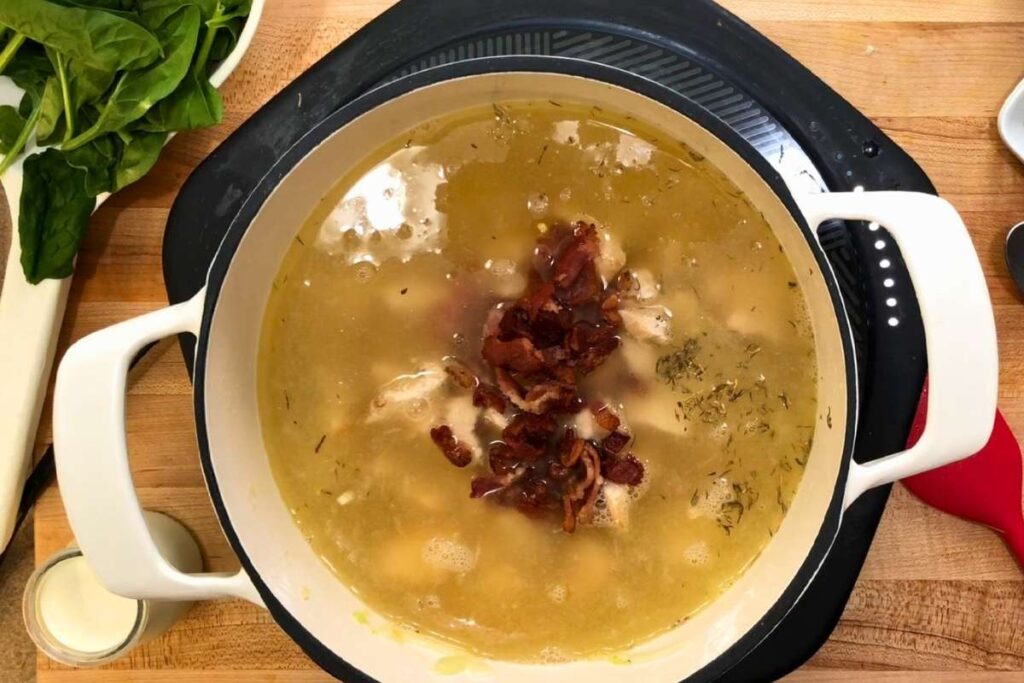 A white pot on a stovetop contains soup with visible herbs and pieces of bacon in broth. Spinach leaves, a cup of cream, and other ingredients are nearby on a wooden counter.