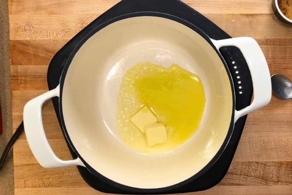 A white pot on a stovetop contains melting butter and oil, sitting on a wooden countertop with a spoon and a small bowl nearby.