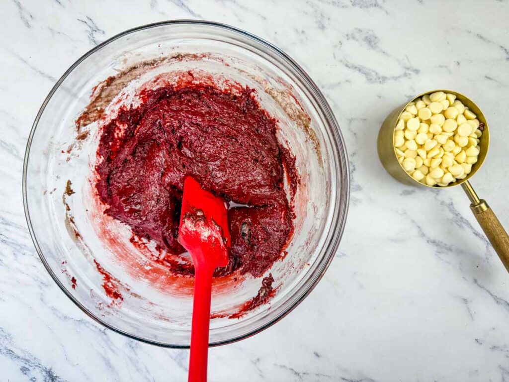 A glass bowl with red cookie dough and a red spatula sits on a marble surface next to a measuring cup filled with white chocolate chips.