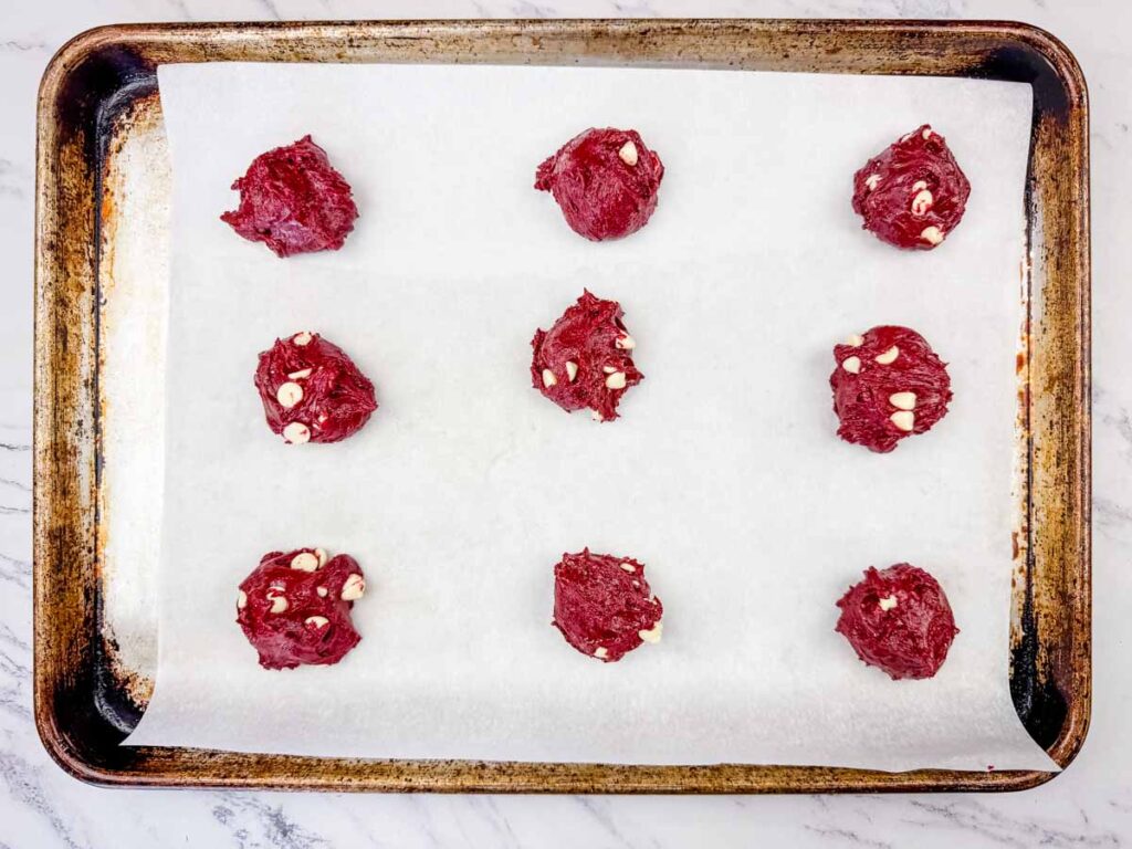 Nine scoops of red cookie dough with white chocolate chips are evenly spaced on a parchment-lined baking sheet.
