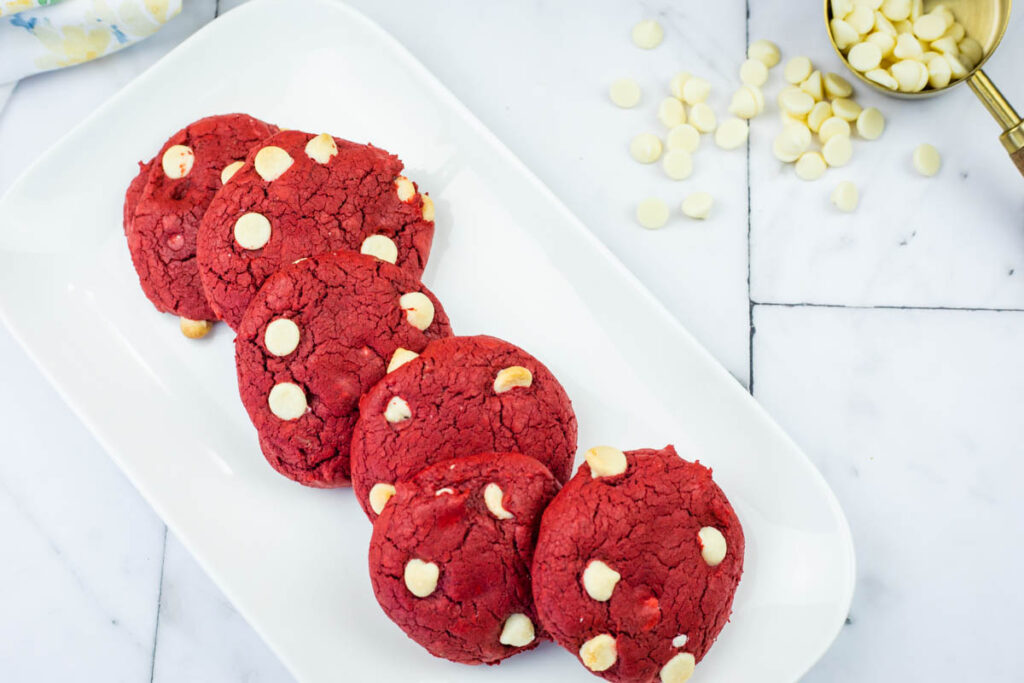 Five red cookies with white chocolate chips arranged on a white rectangular plate, with extra white chocolate chips scattered nearby on a marble surface.