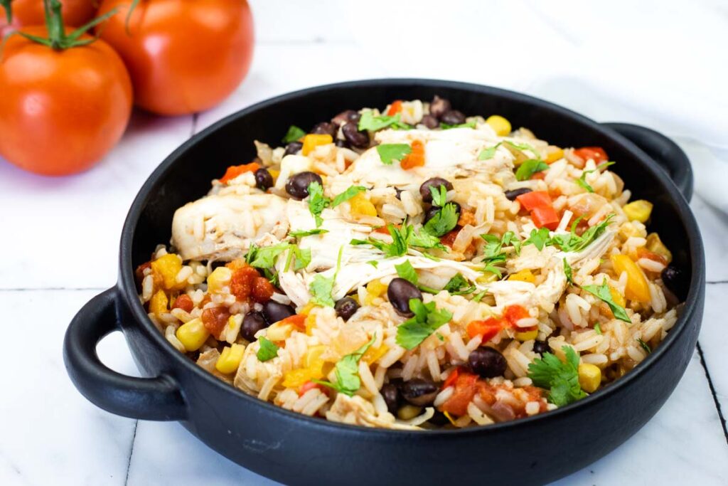 A black skillet filled with rice, black beans, corn, tomatoes, shredded chicken, and chopped cilantro on a white surface. Three whole tomatoes are in the background.