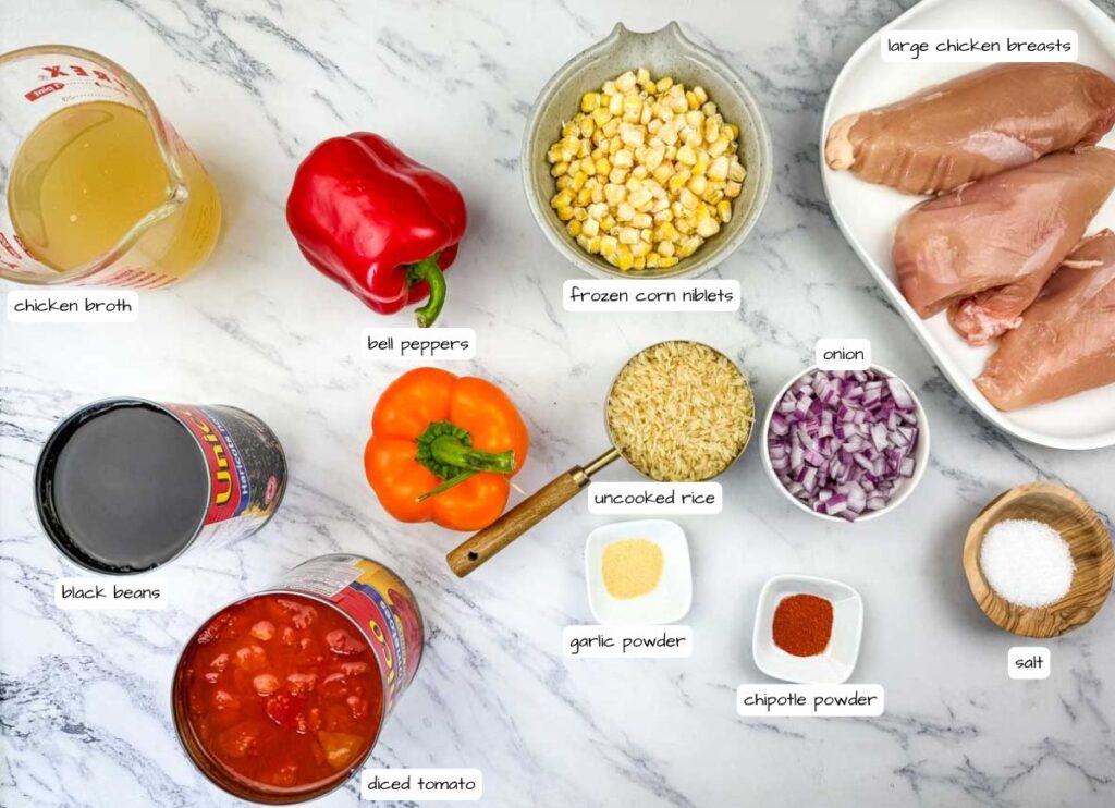 Ingredients for a chicken rice dish arranged on a marble surface, including chicken breasts, rice, broth, corn, bell peppers, onion, black beans, diced tomato, and seasonings.