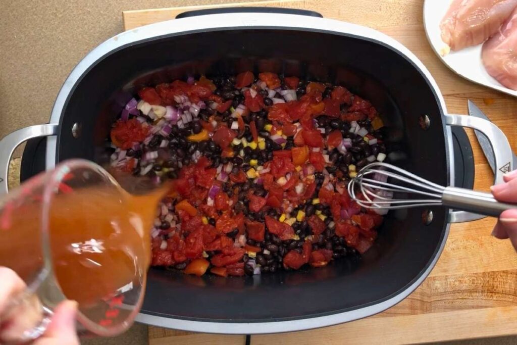 A slow cooker filled with beans, corn, onions, and tomatoes, with broth being poured in and a whisk resting inside; raw chicken breasts are on a plate nearby.